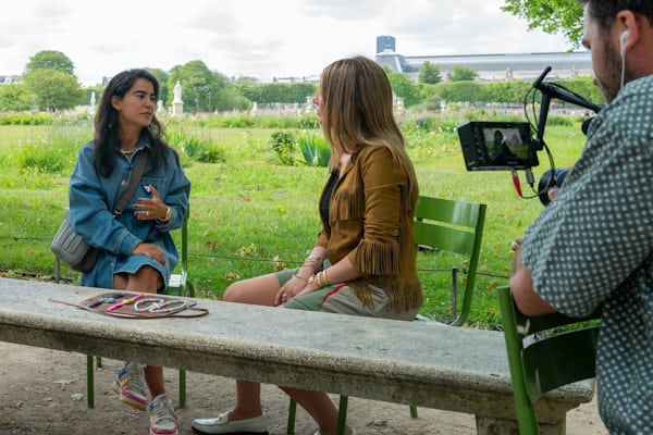 Malaika Crawford and Nadine Ghosn sit and talk at a table in an outdoor park with watches on display while a cameraman films their conversation