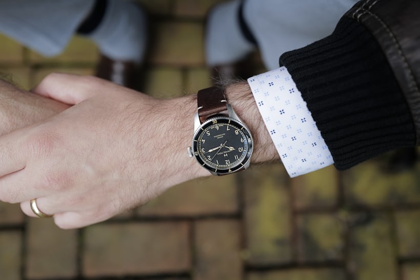 The Hamilton Khaki Pilot Pioneer in 38mm size on wrist with hands crossed and brown leather jacket.