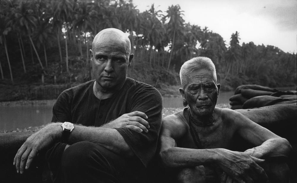 Two actors sit next to each other outdoors in a scene from Apocalypse Now