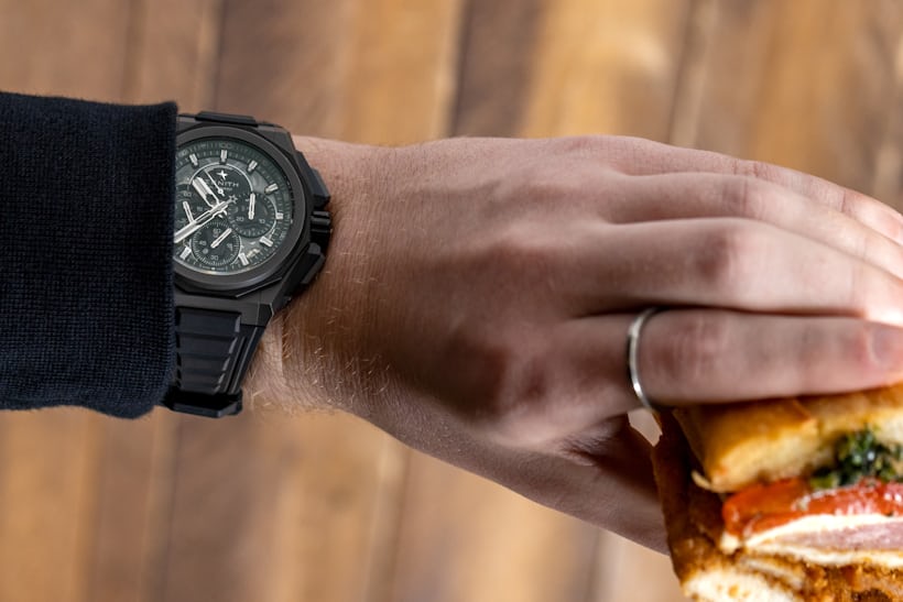 A wrist shot of a man wearing a Zenith Defy Extreme and holding a sandwich in his hand. 