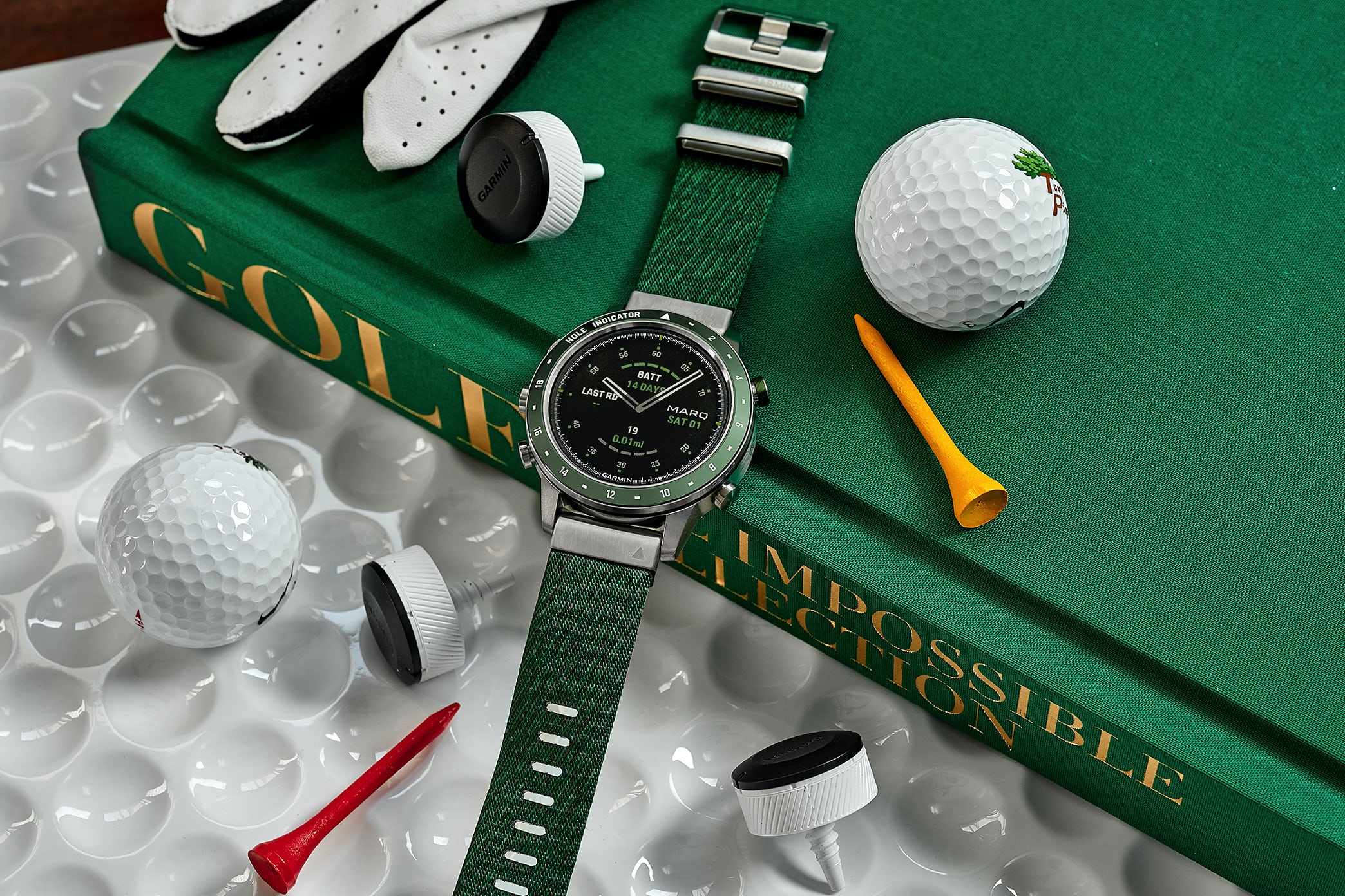 The Garmin MARQ Golfer 46mm laying atop Assouline's "Golf: The Impossible Collection" with assorted golf paraphernalia laying around it.