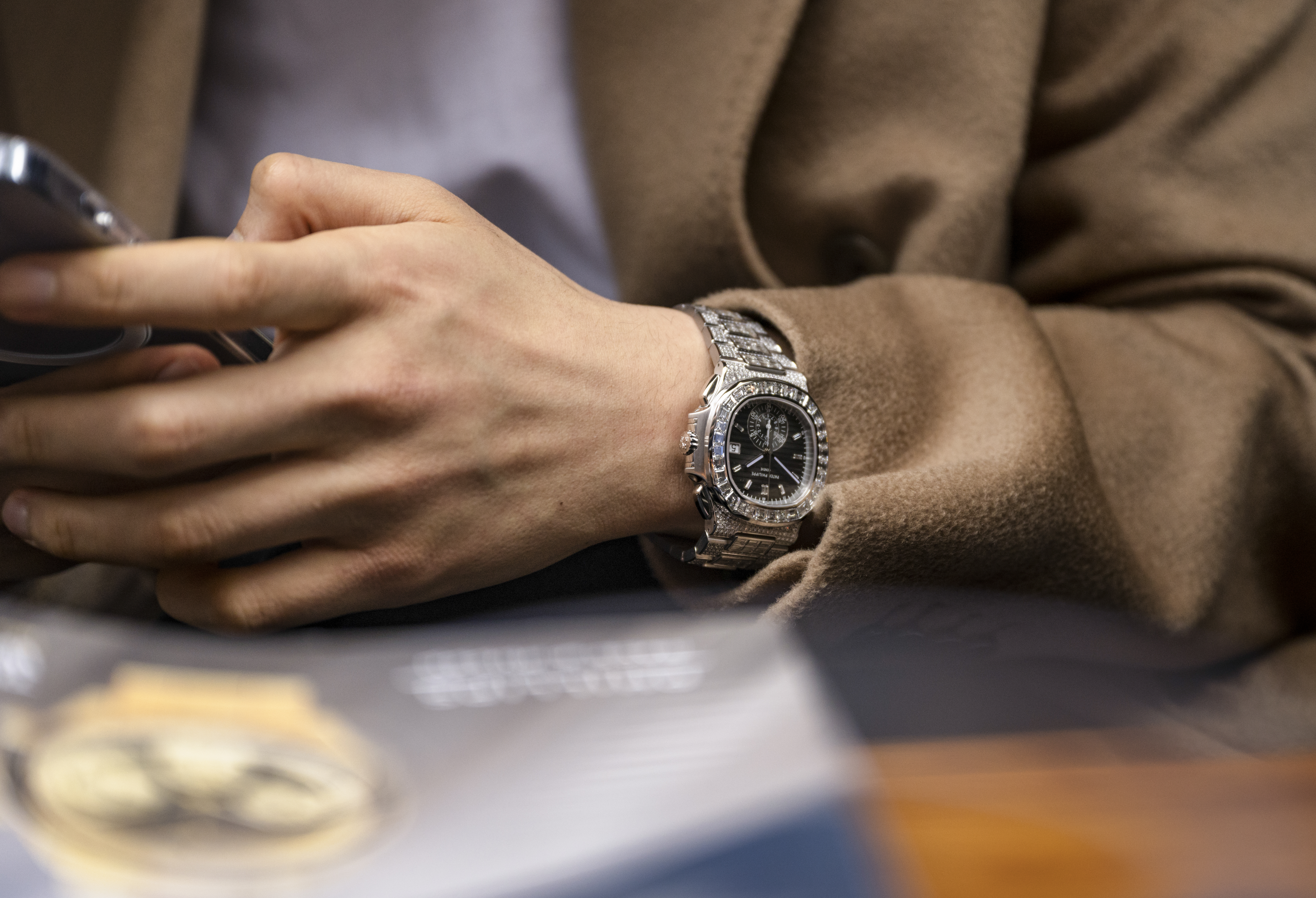 A man in a tan coat wears a Patek Philippe Nautilus 5980-1400G