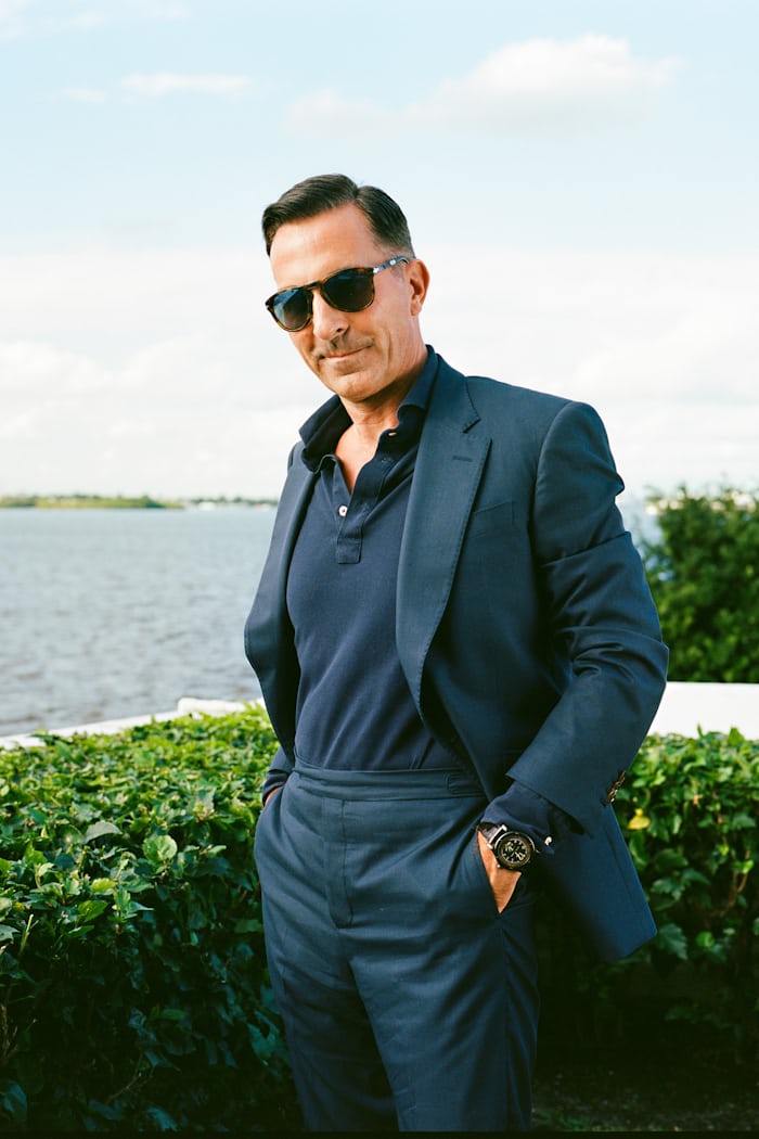 A man wearing sunglasses and a suit poses in front of water