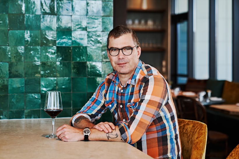 Hugh Acheson sits at a table with a glass of wine 