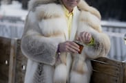A person in a white fur coat leaning against a fence