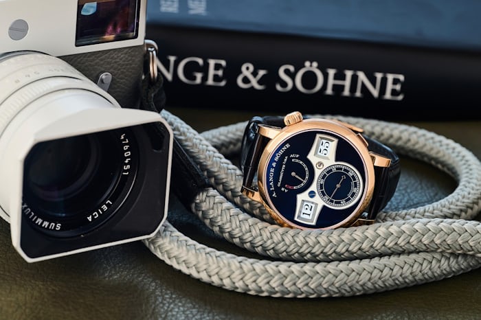 The A. Lange & Söhne Zeitwerk watch sitting on a camera strap next to a Leica camera and a book