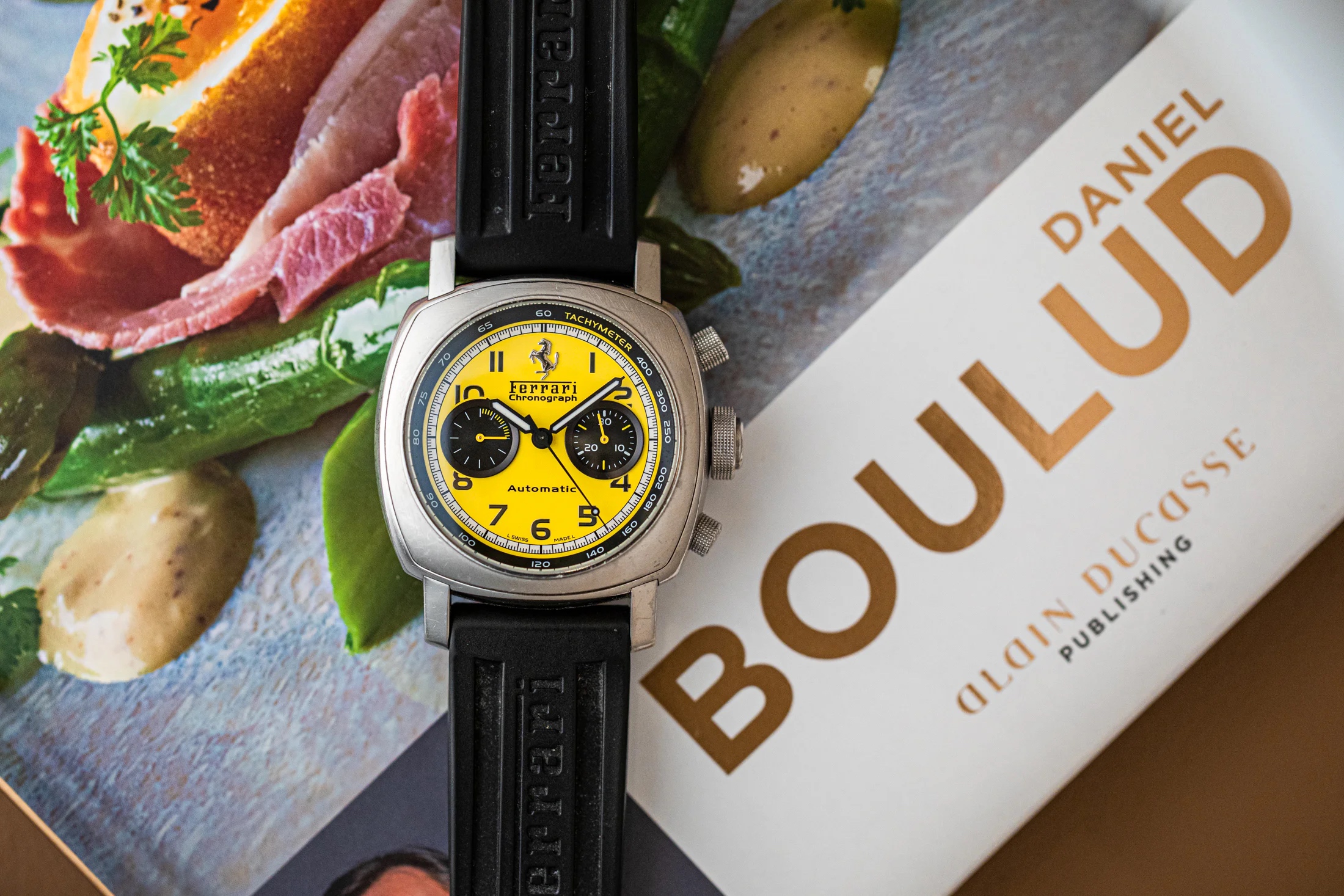 Yellow Ferrari Chronograph positioned on top of a book page featuring colorful food and the name "DANIEL BOULUD"