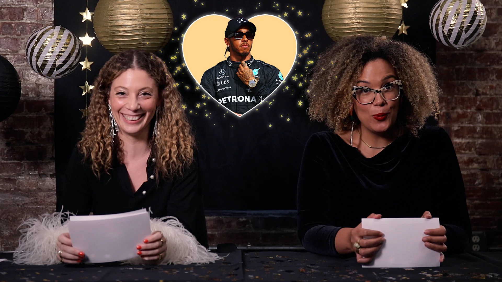 Malaika Crawford sits to the left of Nora Taylor at a table decorated with New Years Eve/Day decor. They are both holding note cards and wear interested expressions. An image of Lewis Hamilton in a glowing heart floats between them