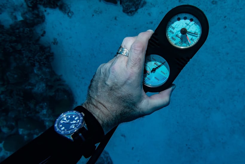 A man wears the Tudor Pelagos FXD underwater and holds a mechanical depth gauge