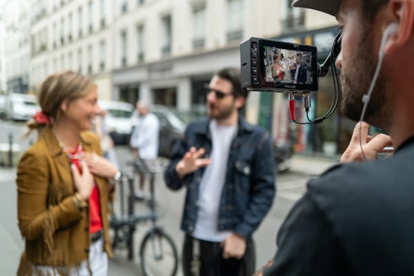 A videographer shoots video of Malaika speaking to a man on the street. Malaika and the man are blurred and in the background while the videographer is in focus in the foreground