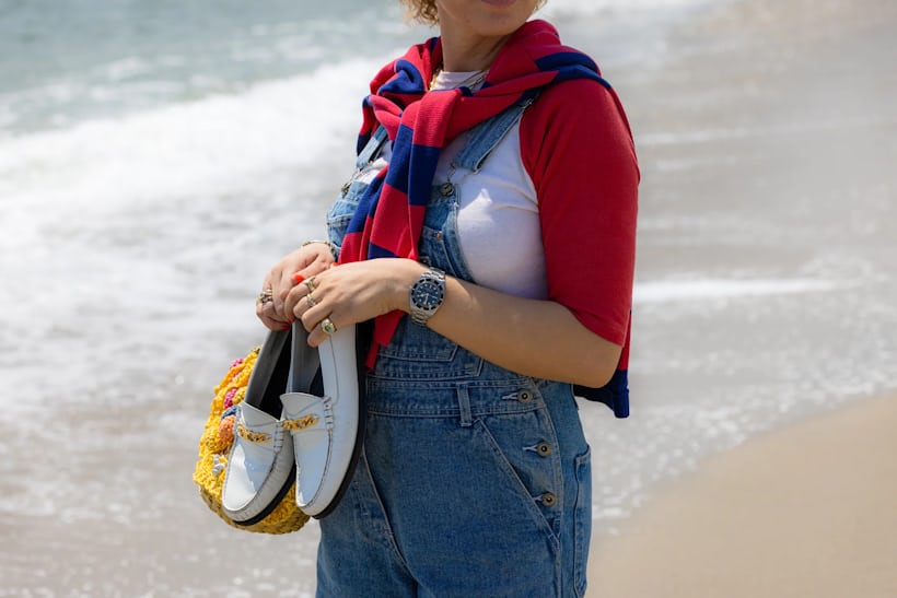 首から太ももまでを切り取った女性の写真。白い靴を履き、チューダー　ブラックベイ 54を手首につけ、潮が満ちてくる砂浜に立っている