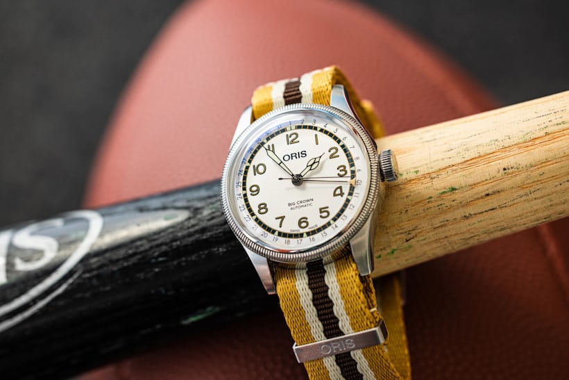 An Oris Big Crown watch is wrapped around a wooden baseball bat resting on top of a football