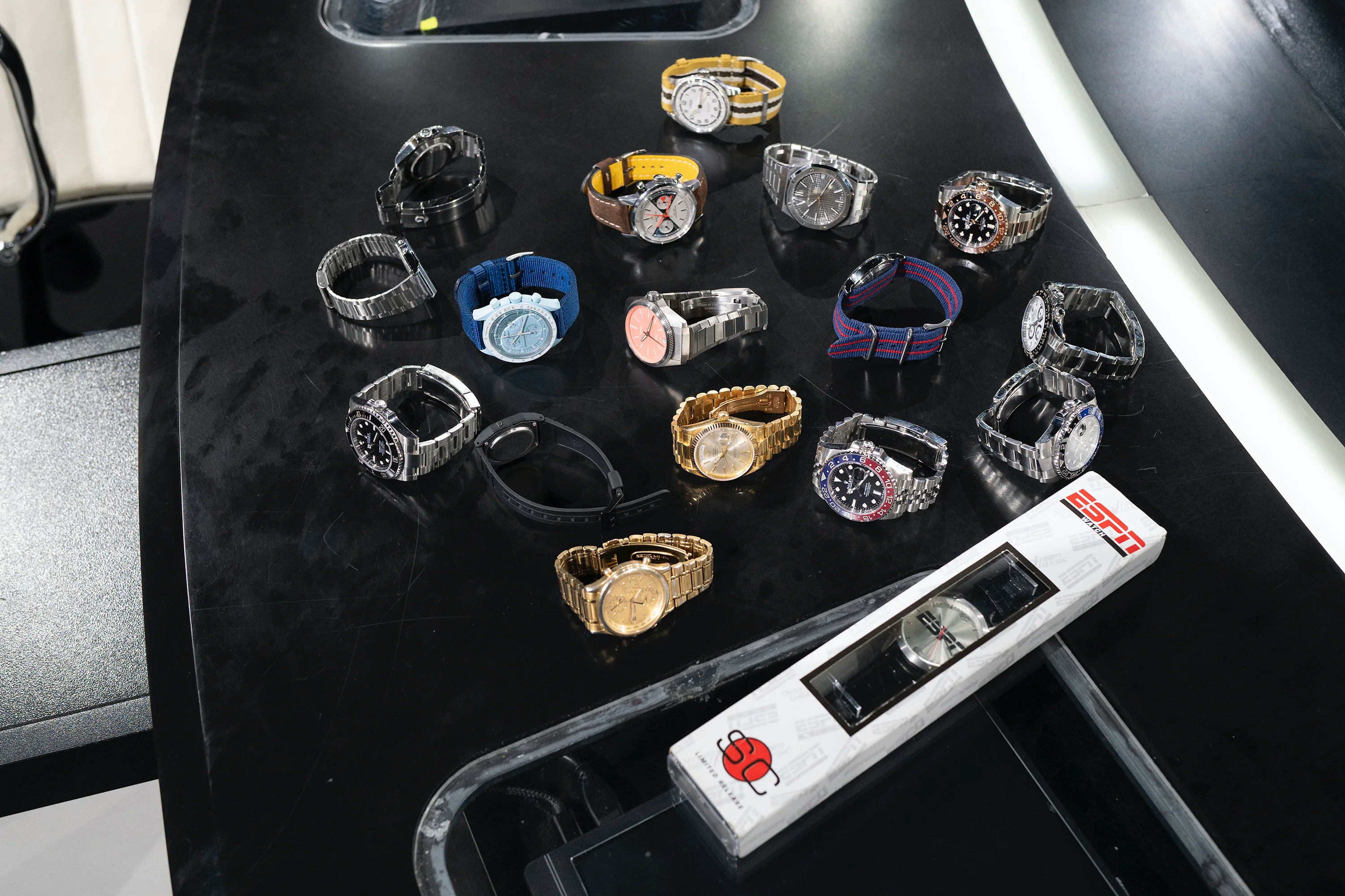 A collection of brand-name watches of varying sizes and colors is laid out on a curved black desk