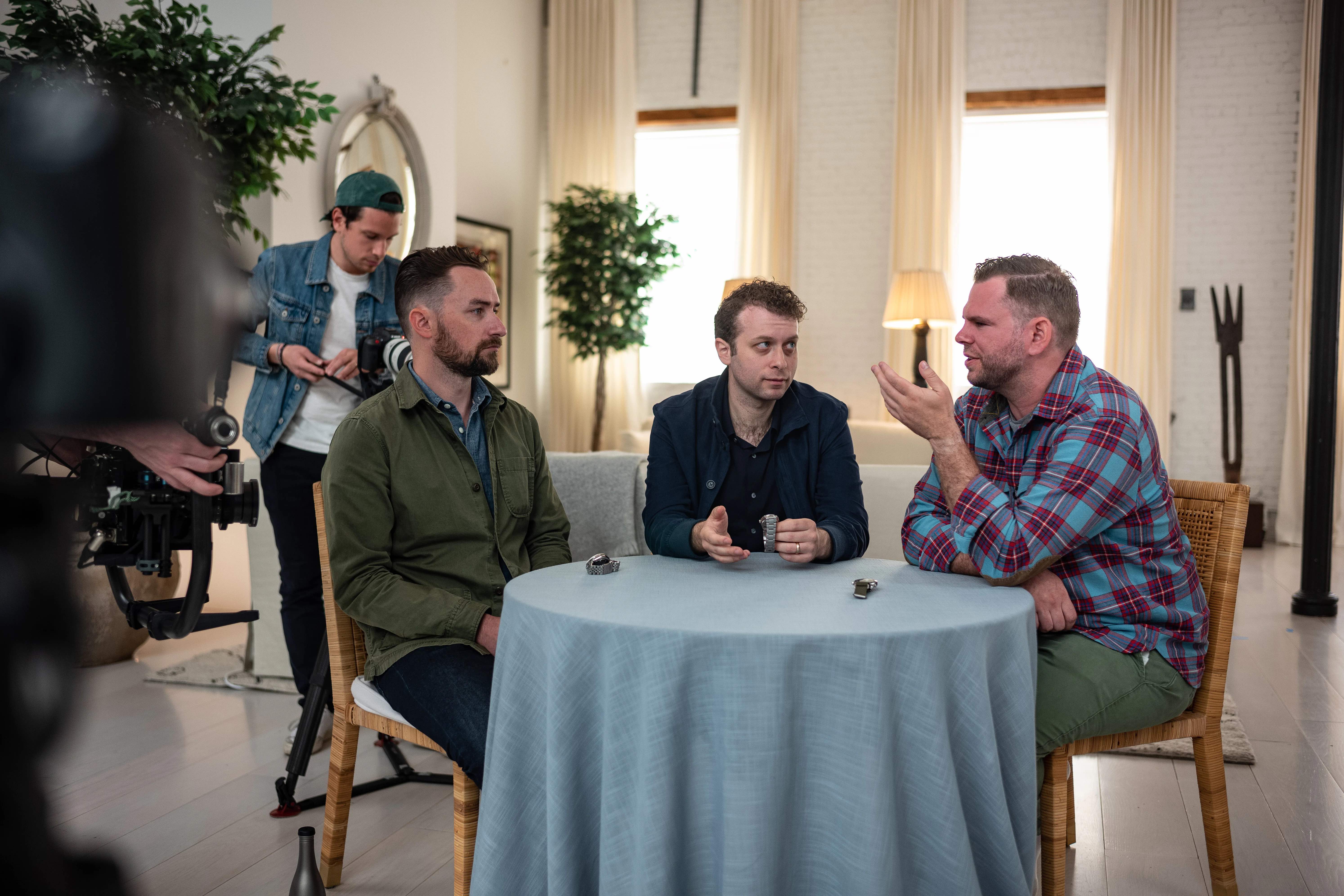 three men sitting around a table, talking with cameraman in the background